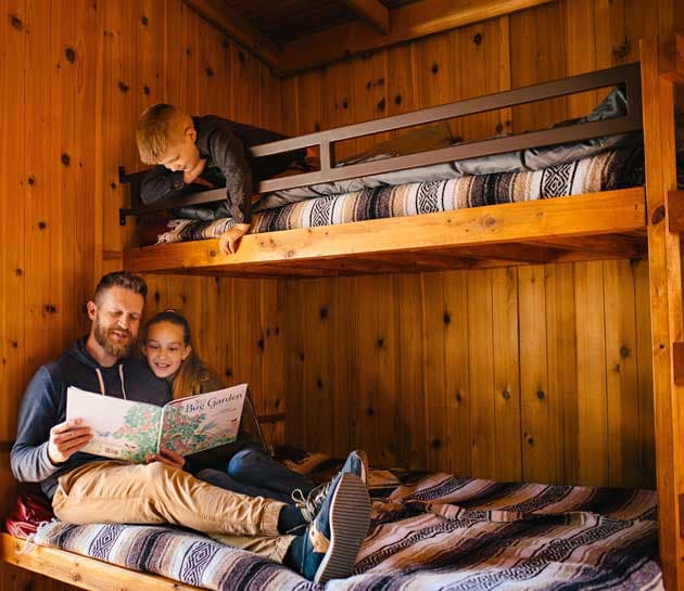 A father reading a book to his 2 children in a bunk room of a cabin at Harts Camp
