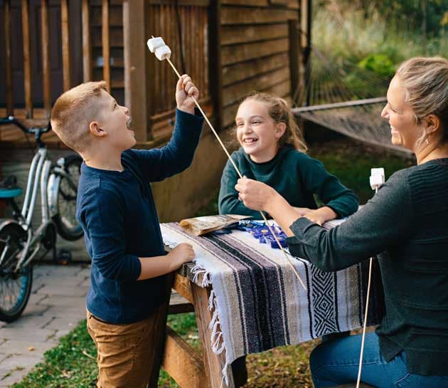 A mother and two children putting marshmallows on sticks outside of the cabin at Harts Camps