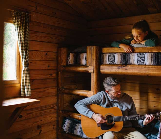 A father playing guitar on the bottom bunk for his daughter in a cabin at Harts Camp