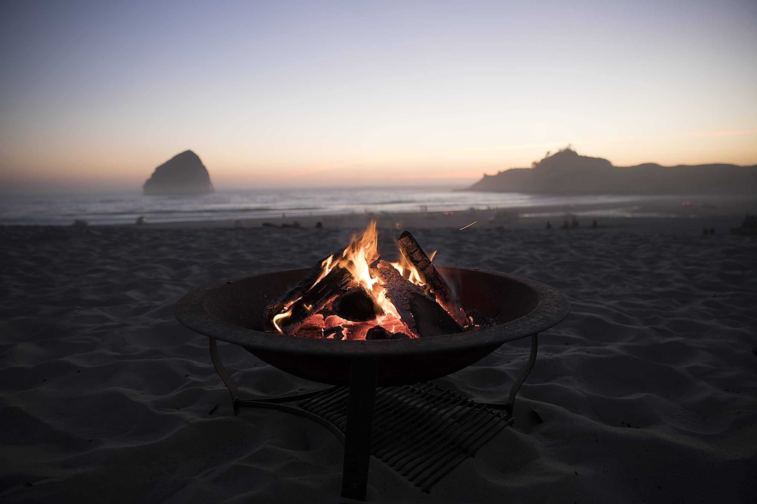 Beach fire pit glowing at sunset on Cape Kiwanda sand with Haystack Rock silhouetted offshore near The Inn at Hart's Camp glamping resort in Pacific City, Oregon Coast.