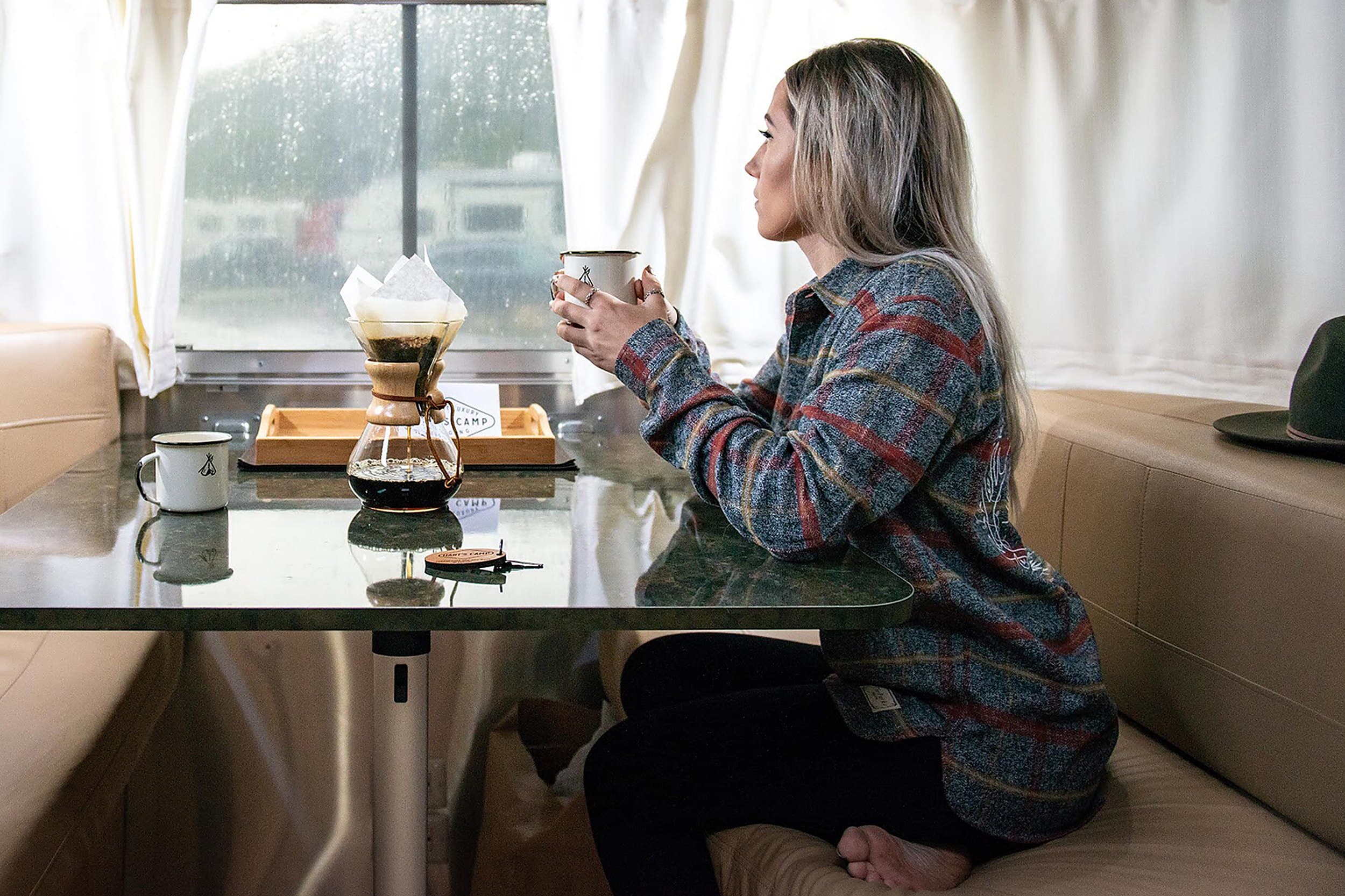 Woman in flannel sipping pour-over coffee inside a vintage Airstream trailer rental at Hart's Camp Pacific City Oregon