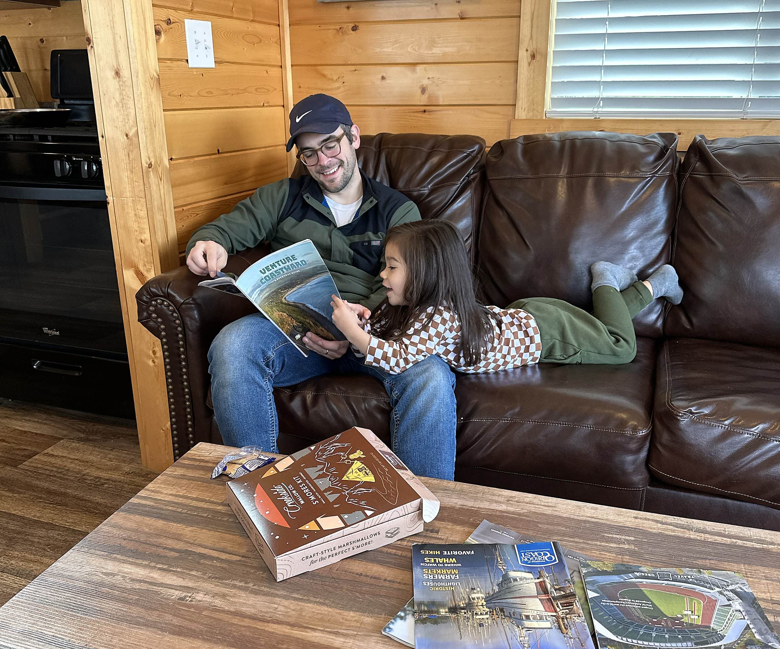 Father and daughter reading an Oregon Coast travel guide with a Creekside Mallow Co. s'mores kit inside a coastal cabin at Hart's Camp Pacific City