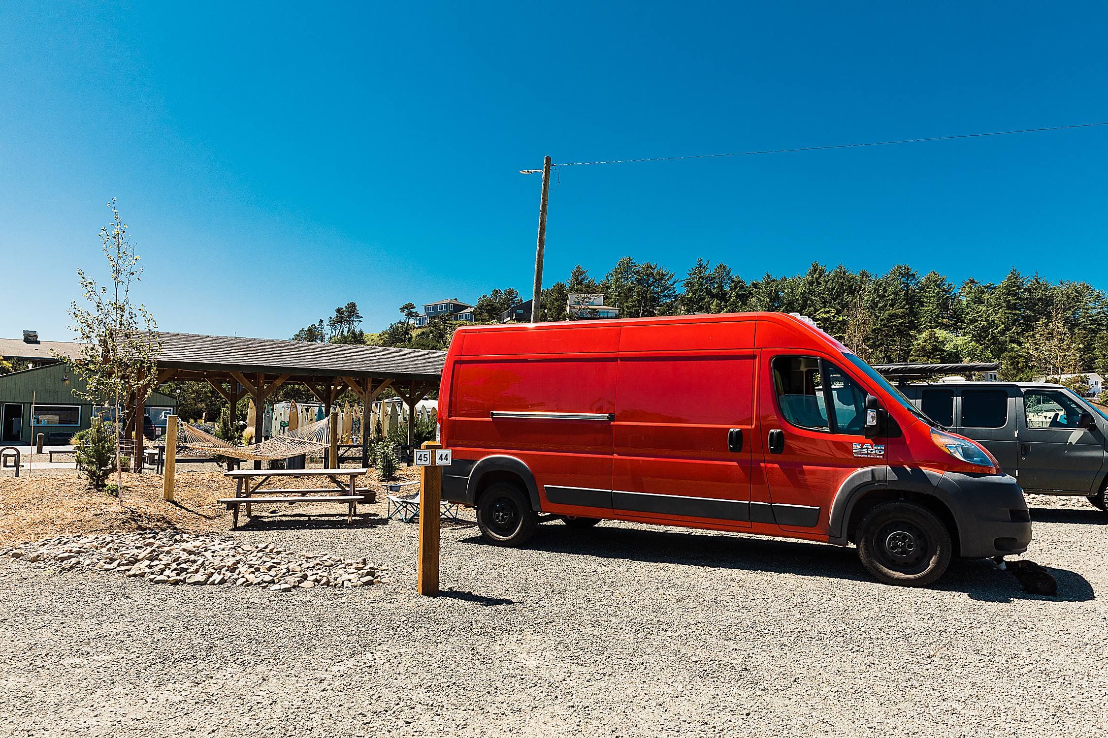 Red Ram ProMaster camper van parked at a sunny RV site with surfboard rack and hammock at The Inn at Hart's Camp in Pacific City, Oregon Coast.
