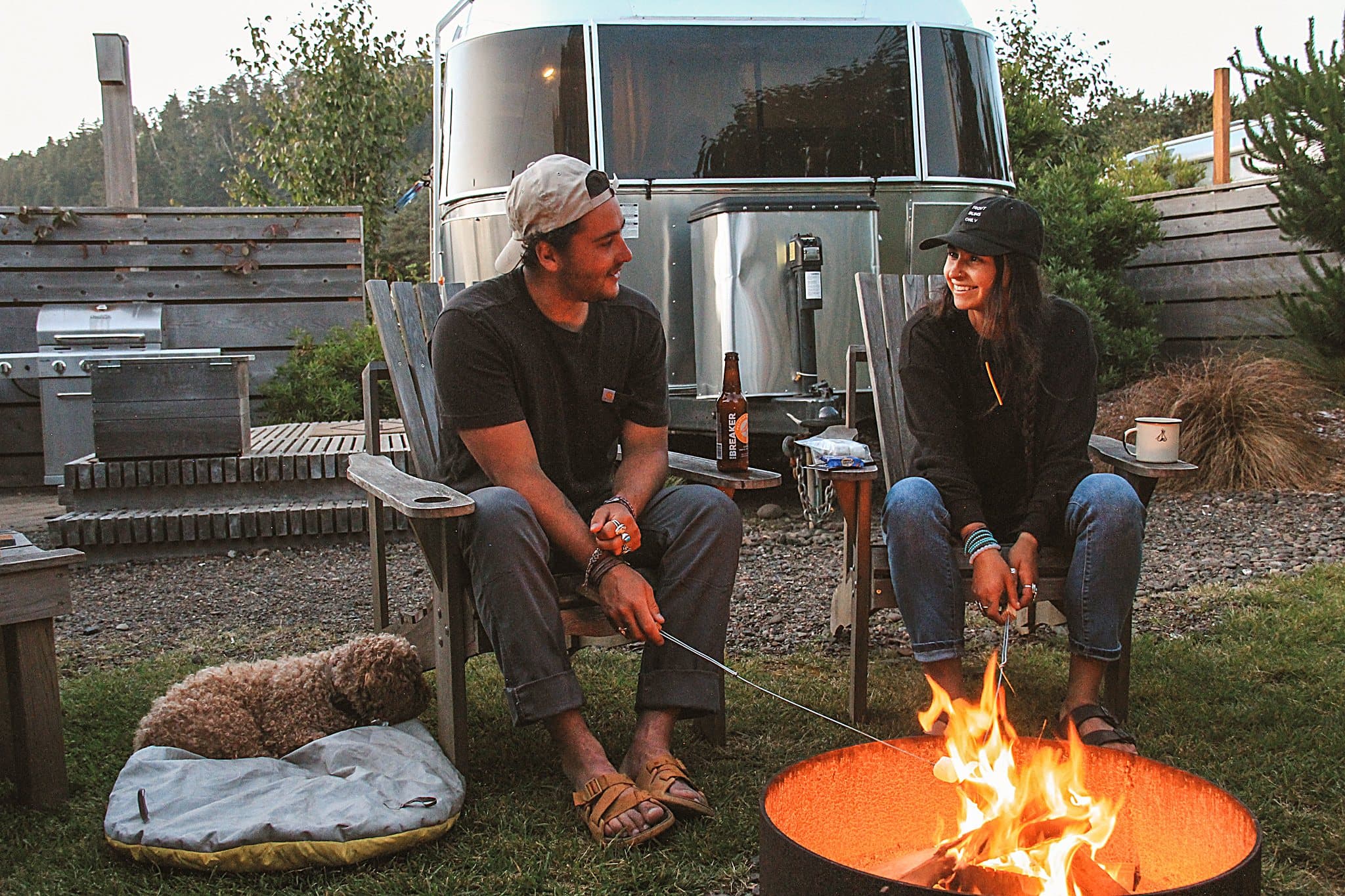 Couple roasting s'mores at a fire pit beside a vintage Airstream at The Inn at Hart's Camp, a glamping resort in Pacific City on the Oregon Coast