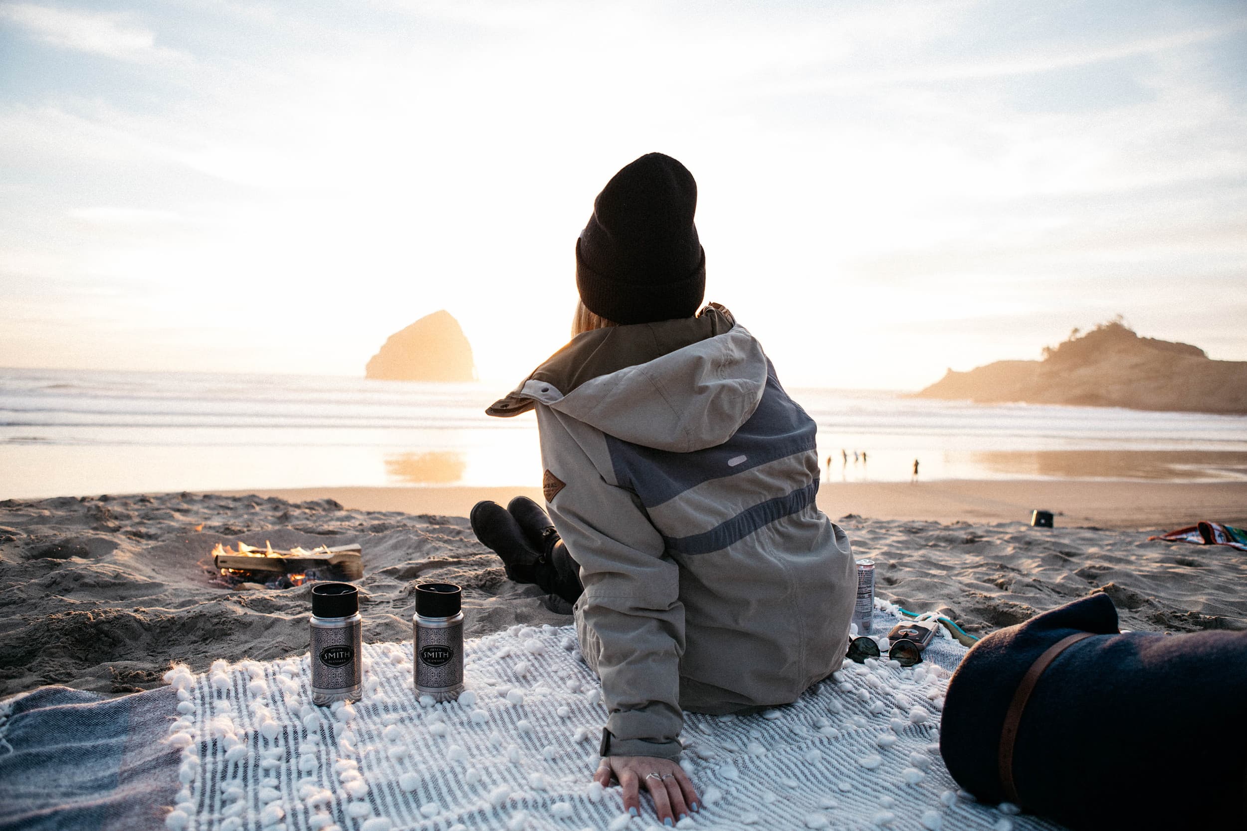 Guest in a beanie and jacket relaxing beside a small beach bonfire at sunset near Hart's Camp glamping resort on the Oregon Coast