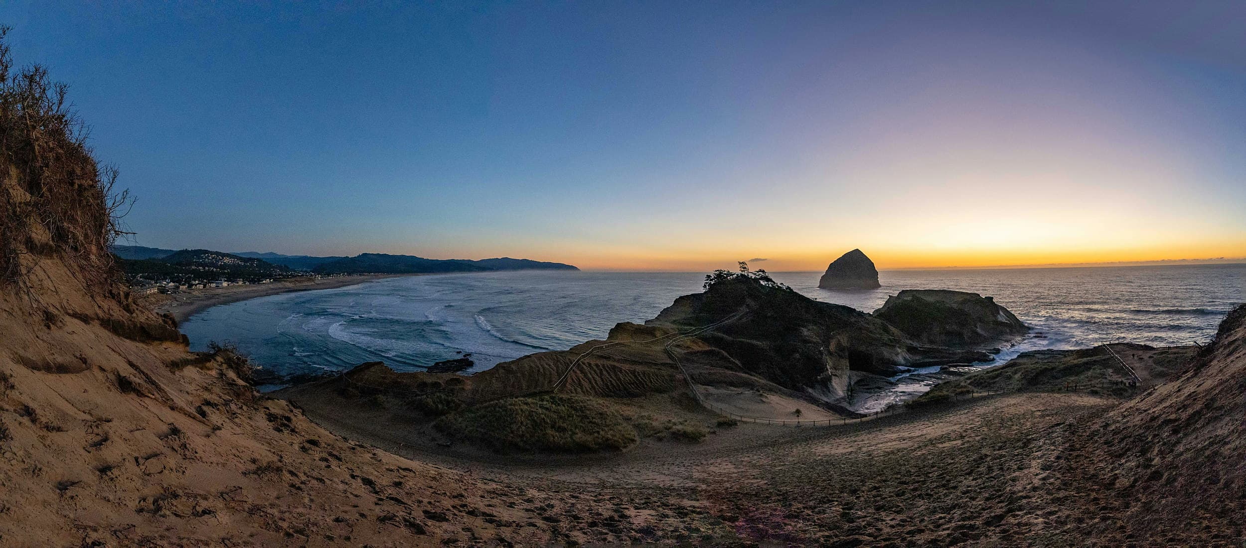 Panoramic view of a sandy cove, headland trail, and offshore sea stack at sunset along a rugged Pacific coastline