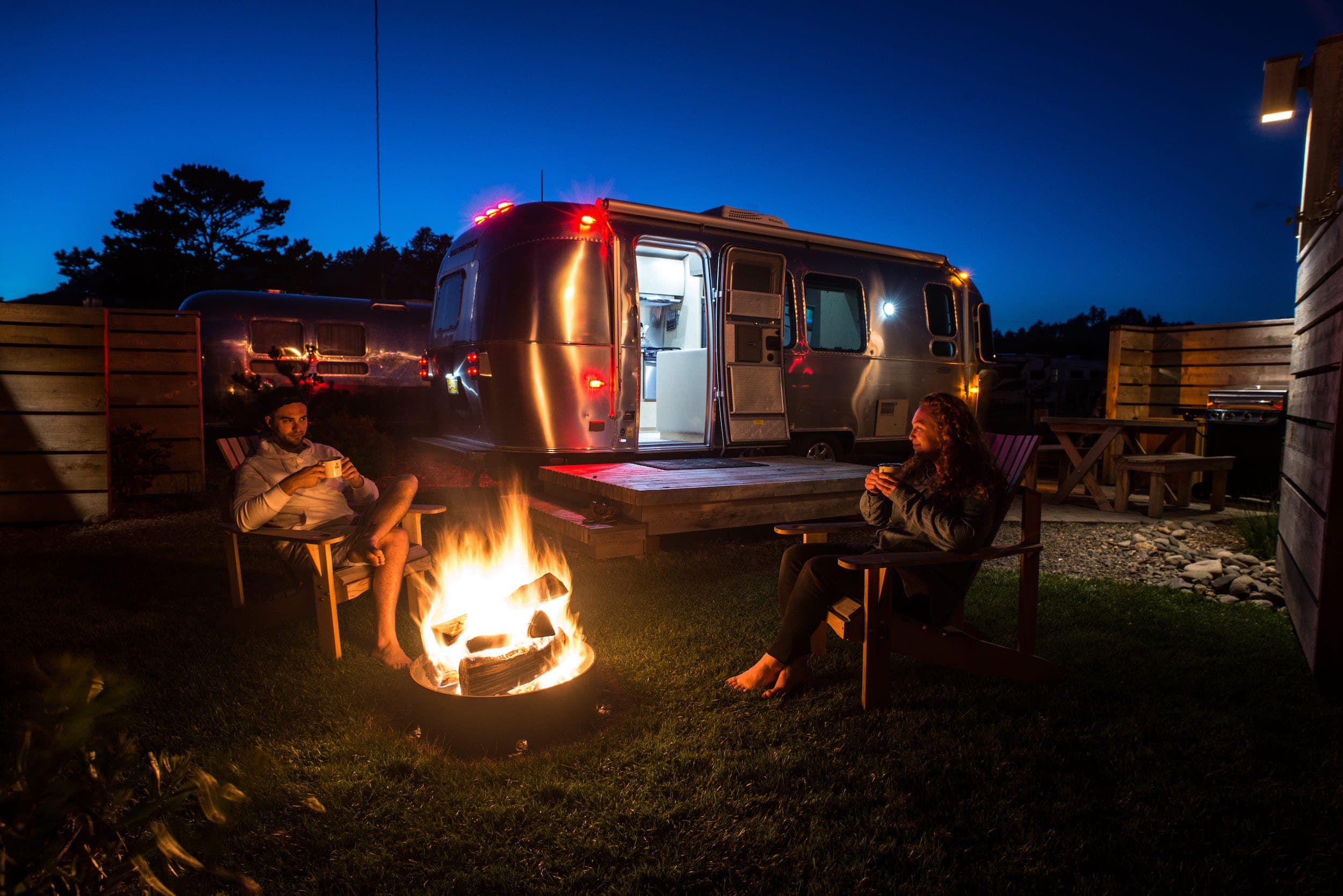 A couple sitting in front of a fire pit outside their Airstream at Harts Camp with warm drinks at night