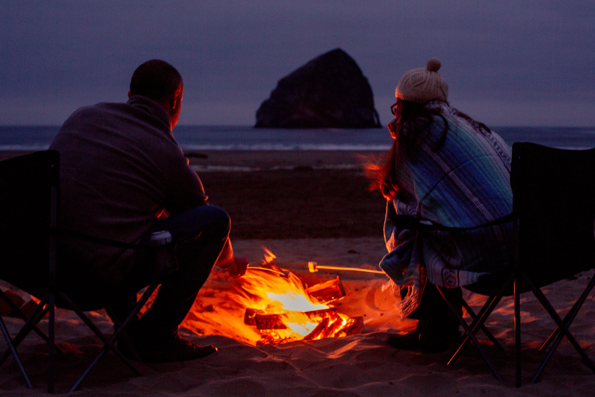 A couple sitting on front of a fire on the beach roasting marshmallows