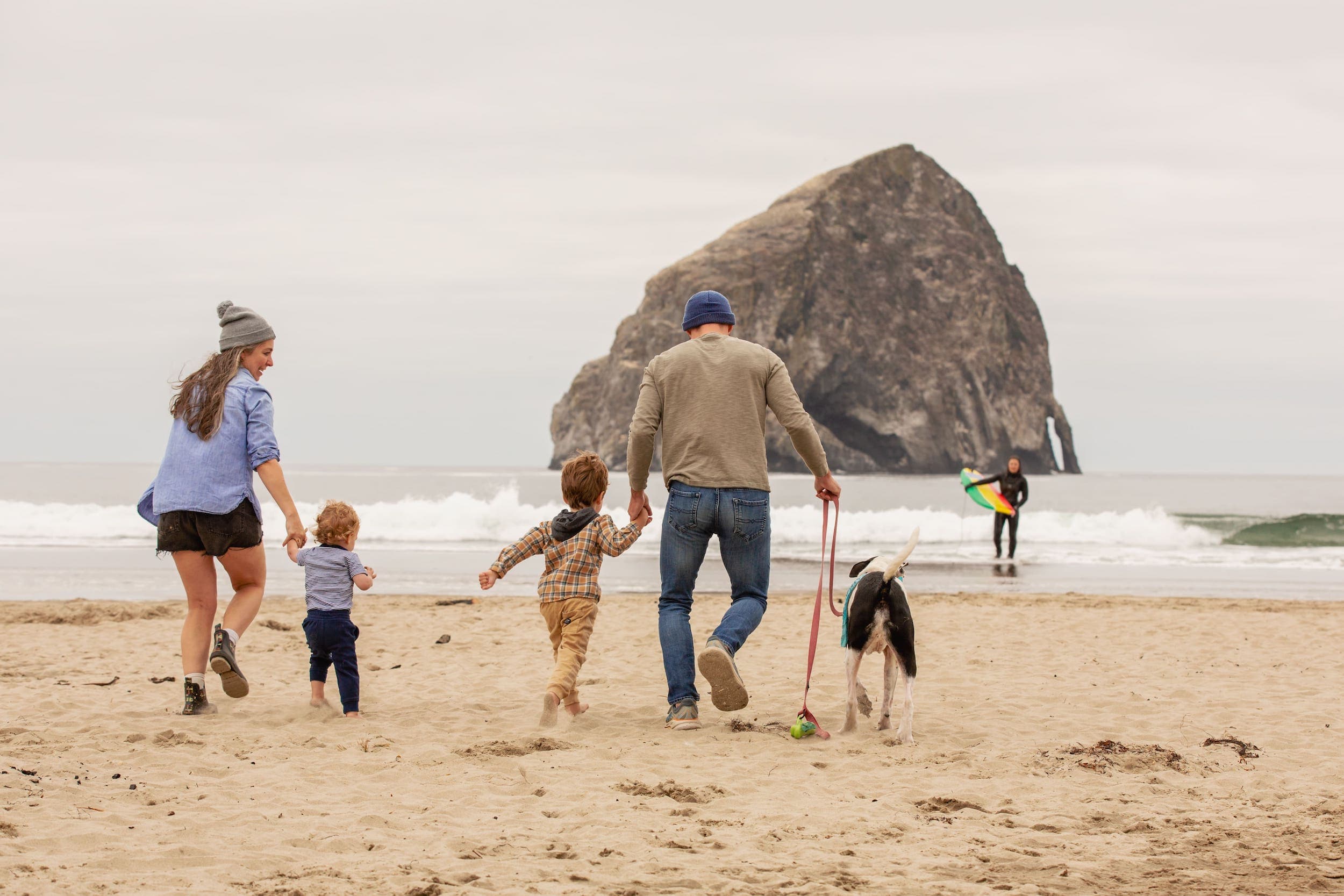 A family walking on the beach with their dog and a surfer holding a surfboard in the shallow water near Hart's Camp
