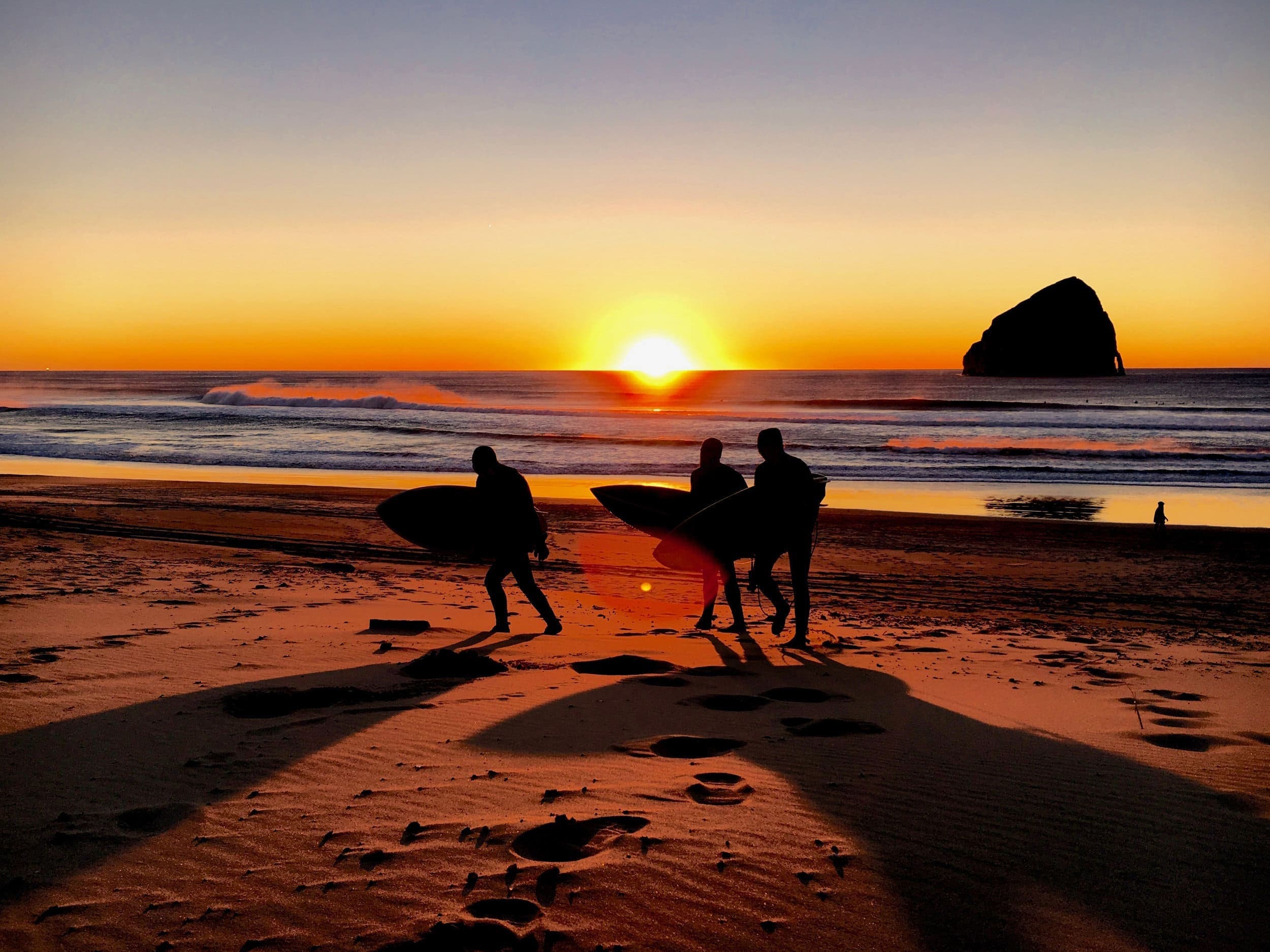 Three surfers walking the shore of the beach as the sun is going down casting beautiful warm yellows and pinks over the ocean water and beach sand