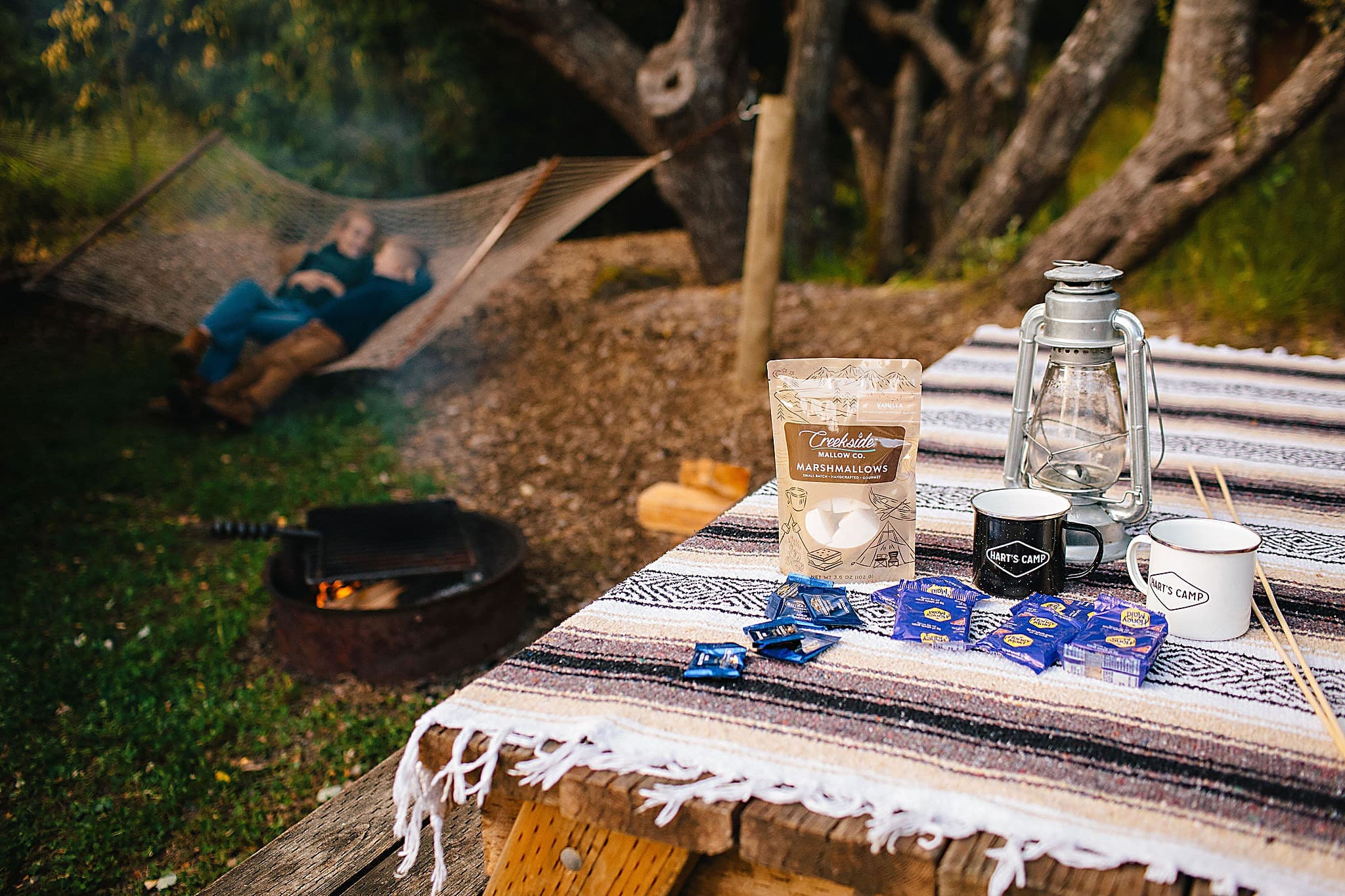 S'mores kit with marshmallows, chocolate, lantern, and Hart's Camp mugs on a picnic table beside a fire pit, with a couple relaxing in a hammock at The Inn at Hart's Camp in Pacific City, Oregon.
