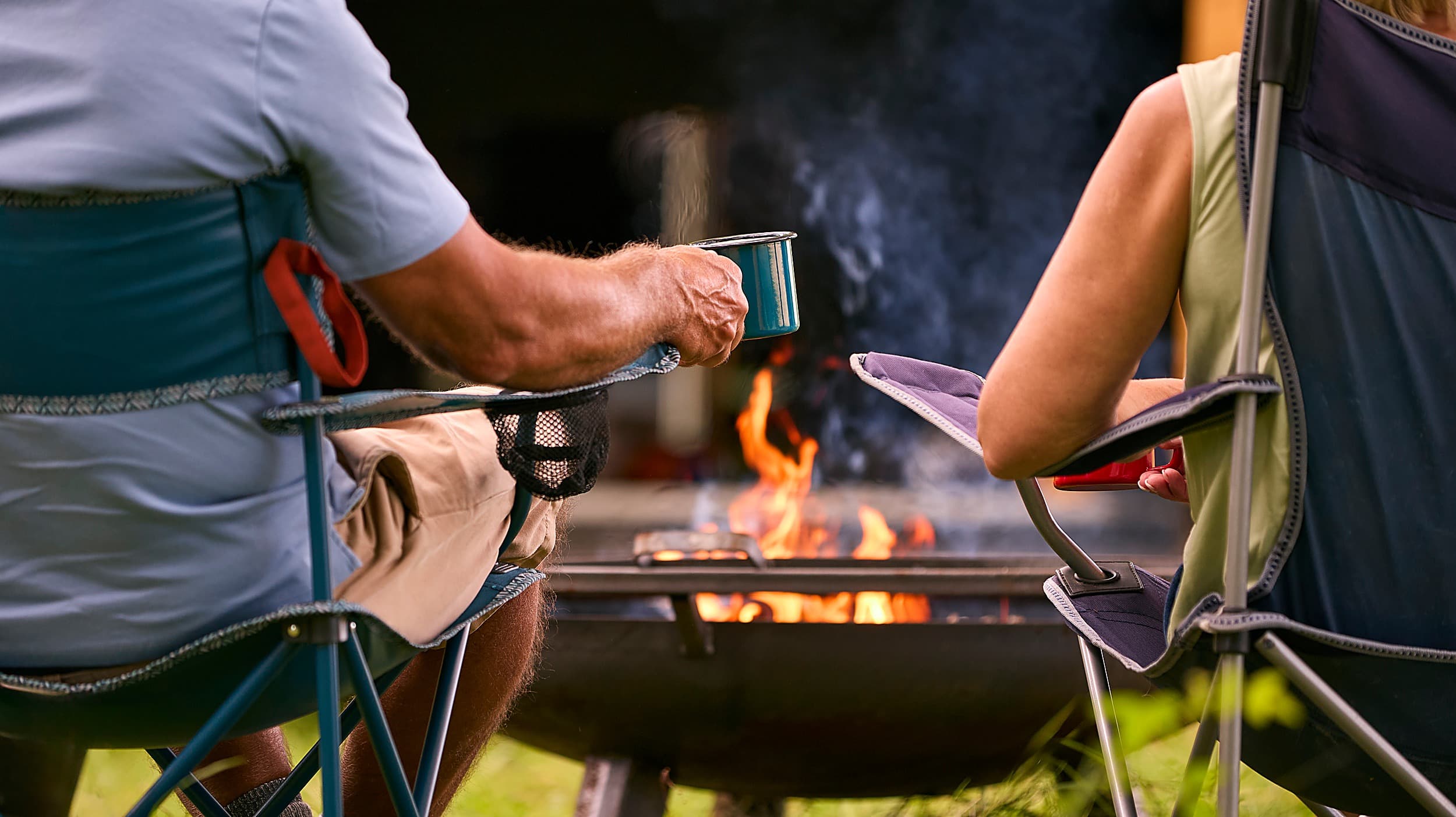 Couple enjoying coffee in camp chairs around an evening campfire at The Inn at Hart's Camp RV resort in Pacific City on the Oregon Coast.