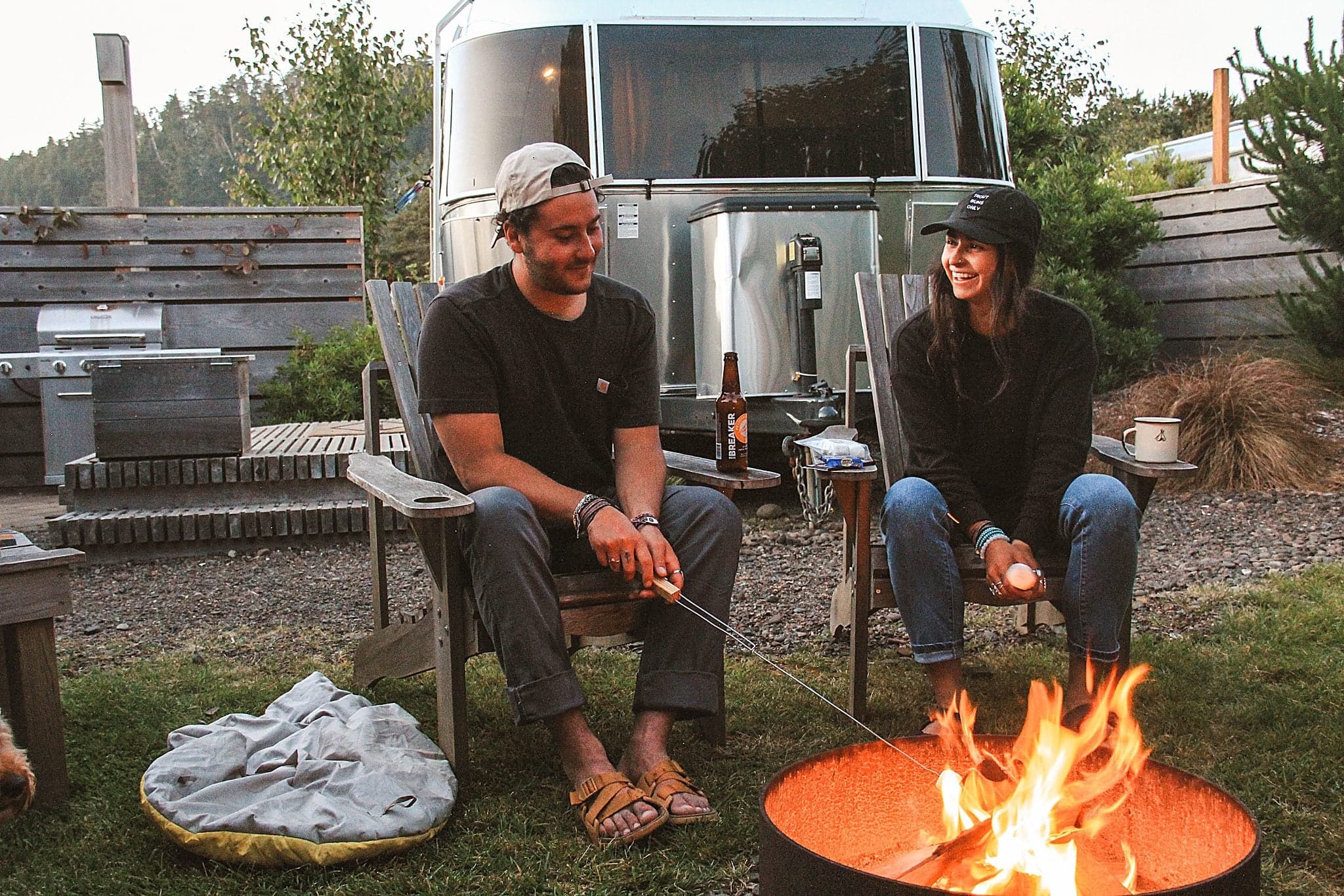 Couple roasting marshmallows at a fire pit beside their Airstream trailer at The Inn at Hart's Camp, a glamping retreat in Pacific City, Oregon.