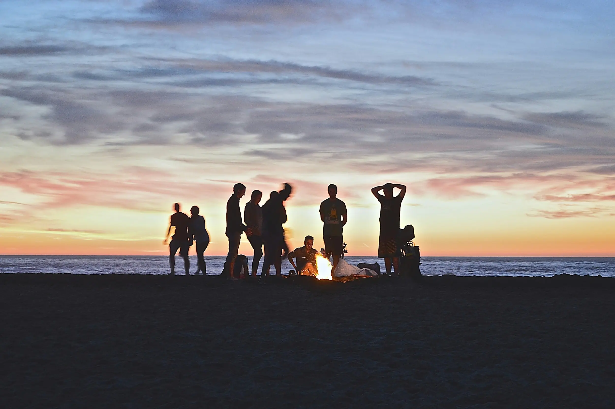 Friends gathered around a beach bonfire at sunset along the Oregon Coast near The Inn at Hart's Camp glamping resort in Pacific City, with Cape Kiwanda and the Pacific surf in the background.